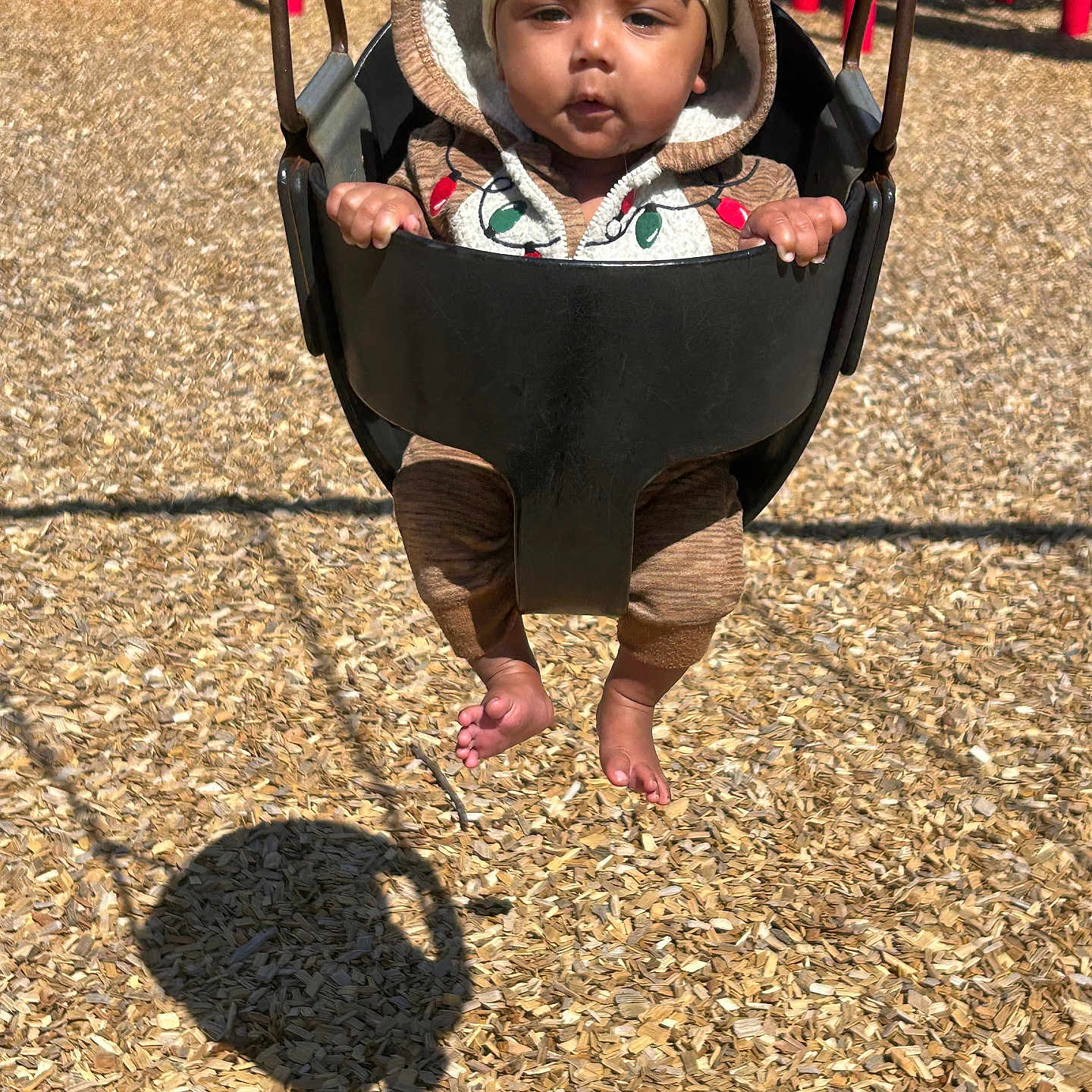 Anna-Mae is registered to the contest to win money with this photo: baby, child, swing, playground, wood_chips, hoodie, headband, yellow, brown, red, grass, sunlight, shadow, outdoor, infant, seat, toy, fun, park, person
