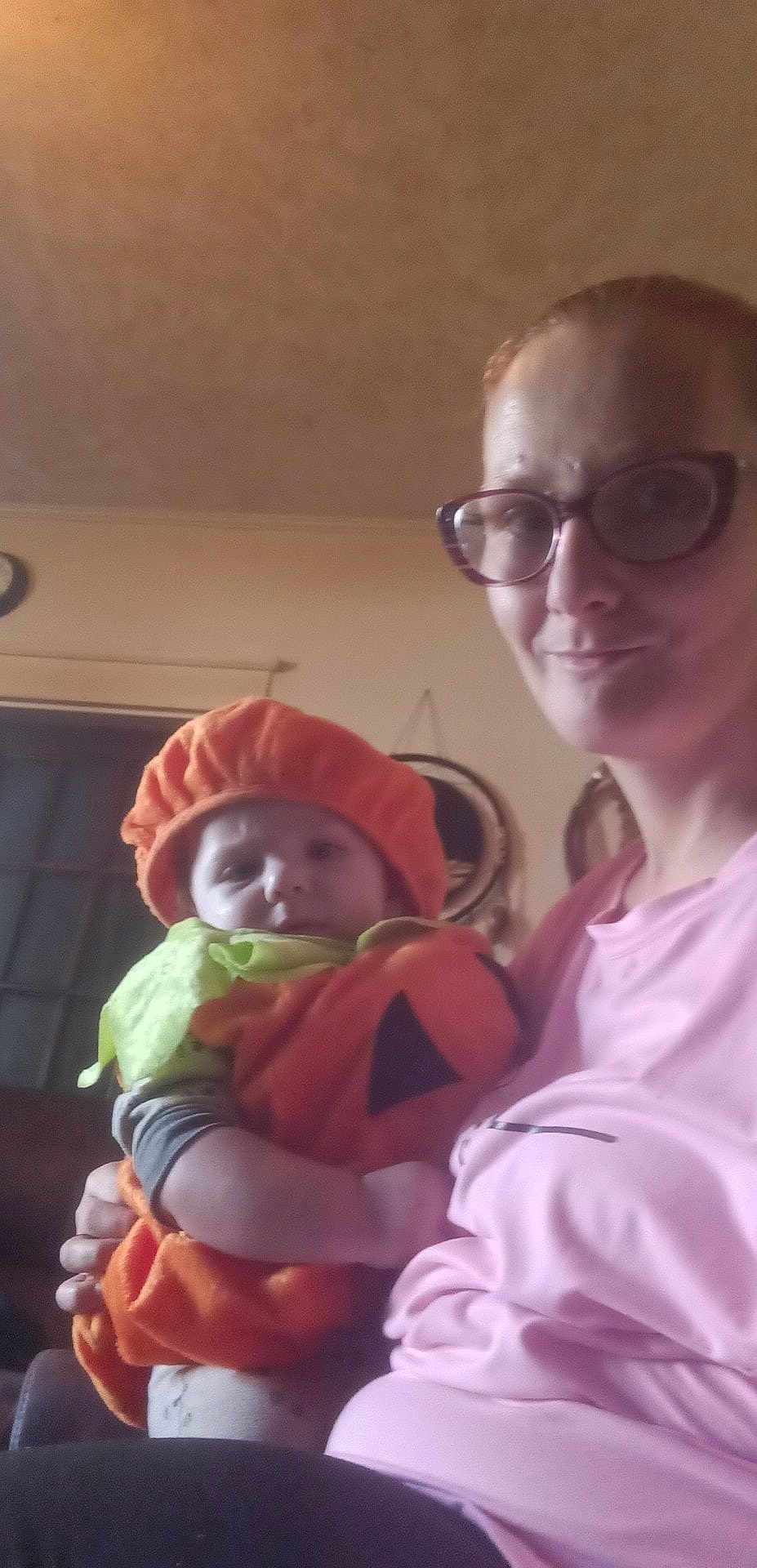 Cole Lee Fowler is registered to the contest to win money with this photo: adult, baby, child, clothing, costume, face, festive, glasses, green, headwear, holding, home, indoor, orange, person, pink, pumpkin_costume, smile, wall, window
