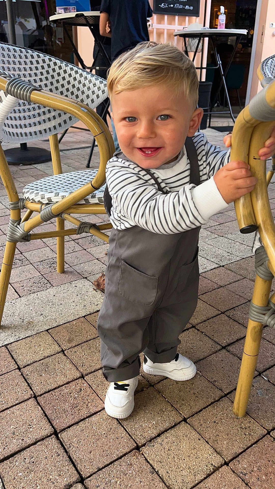 Meylan participe au concours pour gagner de l'argent avec cette photo : toddler, child, smiling, standing, striped_clothing, overalls, white_sneakers, wicker_chair, outdoor, cafe, brick_floor, happy, blue_eyes, short_hair, person, daylight, casual_wear, cute, playful, holding