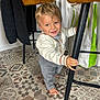 toddler, child, barefoot, indoor, floor, patterned_floor, chair, metal_bar, clothing, smiling, blonde_hair, blue_eyes, person, home, bag, curtain, sweater, pants, furniture, happy