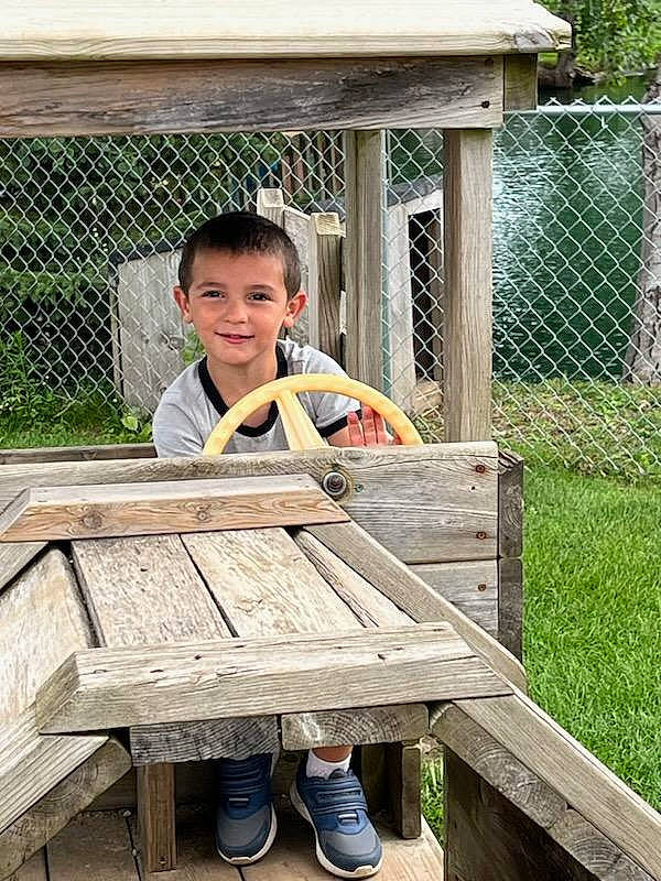 Brodie is registered to the contest to win money with this photo: child, fence, fun, grass, happy, hardwood, joy, leg, leisure, outdoor_bench, outdoor_furniture, person, recreation, shorts, sitting, smile, standing, toddler, vacation, wood