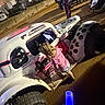 child, race_car, dirt_track, night, people, vehicle, tire, mud, outdoor, curiosity, pink_clothing, shorts, vintage_car, spectators, racing, event, illumination, dirt, automobile, crowd
