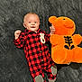 baby, infant, smiling, red_checkered_onesie, plush_toy, orange_teddy_bear, soft_blanket, lying_down, cute, child, happy, cozy, indoor, small_hands, socks, playful, toy, fabric, portrait, young