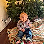 baby, child, plaid_shirt, jeans, barefoot, blanket, christmas_tree, lights, holiday, indoor, floor, wooden_floor, smile, cute, festive, decor, person, sitting, young_child, home