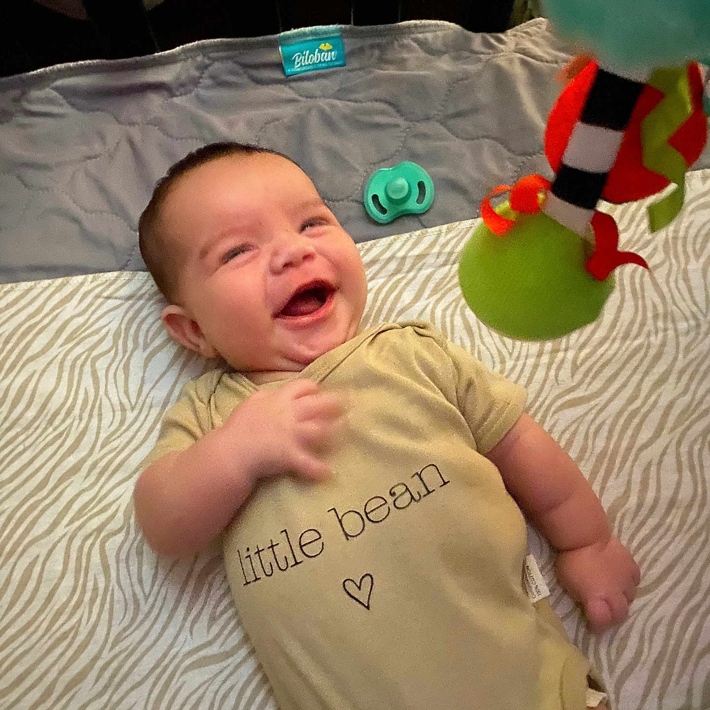 Ryker Zuniga is registered to the contest to win money with this photo: baby, infant, smile, onesie, toy, blanket, pattern, lying_down, happy, cute, child, playful, indoors, face, hand, clothing, pacifier, fabric, colorful, doll