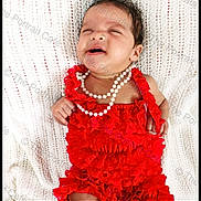 Ariyah is registered to the contest to win money with this photo: baby, infant, red_clothing, pearls, knitted_blanket, cute, portrait, child, smiling, lying_down, indoors, young, adorable, fashion, accessory, happy, skin, hair, expression, closeup