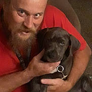 Thor is registered to the contest to win money with this photo: person, beard, dog, puppy, pet, collar, red_shirt, hand, hug, sitting, indoor, couch, portrait, eyes, face, friendship, warm_lighting, closeup, expression, carpet