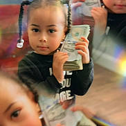 Abrielle is registered to the contest to win money with this photo: blur, braids, cash, casual_clothing, child, closeup, currency, expression, face, hands, holding, indoor, indoor_lighting, money, person, portrait, reflection, stack, wooden_floor, young