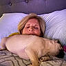 woman, dog, pug, bed, blanket, pillow, indoor, resting, sleeping, pet, headboard, cozy, relaxation, light, fur, comfort, laying, face, human, animal