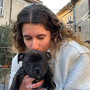 Vyron participe au concours pour gagner de l'argent avec cette photo : person, puppy, dog, holding, kissing, curly_hair, white_coat, ring, outdoor, building, shutters, railing, plant, street, portrait, face, paws, fur, eyes, nose