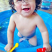Obed is registered to the contest to win money with this photo: toddler, child, curly_hair, water, blue_pool, toy_shovel, yellow_shovel, red_shovel, smiling, happy, playtime, outdoor, water_droplets, bare_chest, splashing, summer, fun, cute, baby, expression