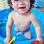 toddler, child, curly_hair, water, blue_pool, toy_shovel, yellow_shovel, red_shovel, smiling, happy, playtime, outdoor, water_droplets, bare_chest, splashing, summer, fun, cute, baby, expression
