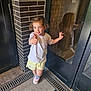 child, girl, smile, thumbs_up, reflection, door, glass, brick_wall, shorts, tshirt, sneakers, blue_eyes, happy, standing, indoor, floor, window, casual_clothing, reflection_in_glass, young