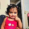 bag, carpet, cheeky_expression, child, curly_hair, cute, doorframe, face, hair_clips, hand, holding, indoor, person, pink_clothing, polka_dot, portrait, smiling, toddler, wall_decoration, young_child
