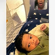 Tavon is registered to the contest to win money with this photo: baby, blanket, clothing, communication, curly_hair, expression, face, green_sweater, home, indoor, infant, lying_down, person, portrait, screen, soft_texture, stars, technology, video_call, yawning