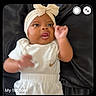 baby, infant, headband, bow, white_clothing, black_background, face, hand, lying_down, curious, expression, text_overlay, video_call, screenshot, person, portrait, indoor, cute, small_face, smiling_adult