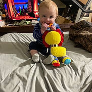 Weston joined the competition — help win amazing prizes! baby, child, toy, plush_toy, bed, blanket, person, indoor, television, woman, curious, chewing, blue_clothing, striped_shirt, sock, furniture, room, home, casual, young_child