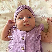 Arabella is registered to the contest to win money with this photo: baby, infant, child, face, headwear, purple_clothing, dress, blanket, pattern, floral, skin, person, cute, lying_down, fist, indoors, portrait, small_child, newborn, soft_texture