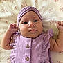 baby, infant, child, face, headwear, purple_clothing, dress, blanket, pattern, floral, skin, person, cute, lying_down, fist, indoors, portrait, small_child, newborn, soft_texture