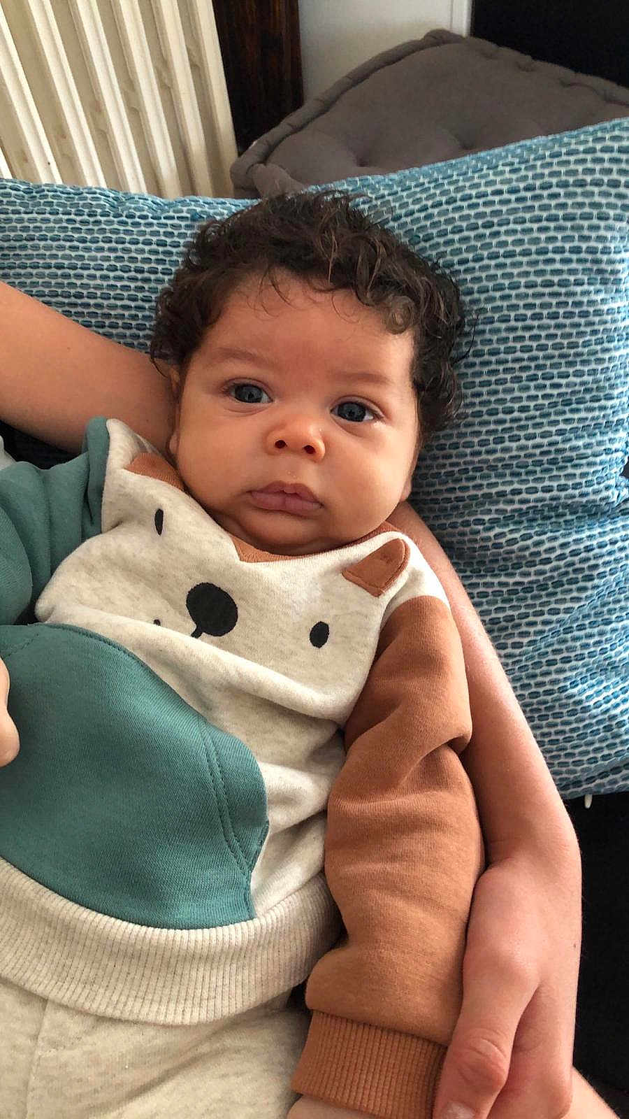 Eden a rejoint le concours — aidez-le/la à gagner de superbes lots ! baby, child, curly_hair, blue_cushion, bear_design, clothing, cute, face, hand, infant, person, portrait, resting, sitting, skin, sleeve, textile, warm_clothing, young_child, indoors