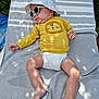 baby, child, diaper, sunglasses, hat, yellow_shirt, lion_design, blanket, outdoor, sunlight, shadow, relaxing, skin, legs, feet, headwear, infant, summer, casual, nature