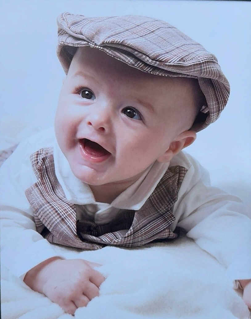 Tommy-Lee is registered to the contest to win money with this photo: baby, infant, smile, cap, plaid, vest, white_blanket, happy, cute, child, portrait, closeup, face, young, clothing, headwear, lying_down, soft_texture, indoor, bright