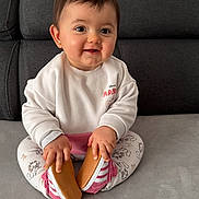 Kayna participe au concours pour gagner de l'argent avec cette photo : baby, infant, child, sitting, smiling, couch, sofa, gray_couch, white_sweatshirt, patterned_leggings, pink_shoes, holding_shoes, hands, face, eyes, hair, earring, home_interior, portrait, soft_lighting