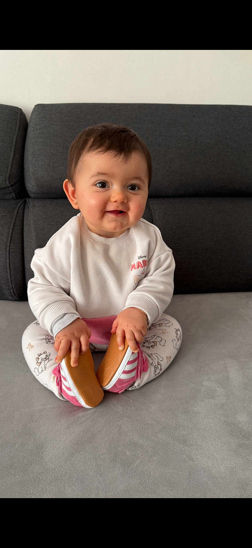 Kayna participe au concours pour gagner de l'argent avec cette photo : baby, infant, child, sitting, smiling, couch, sofa, gray_couch, white_sweatshirt, patterned_leggings, pink_shoes, holding_shoes, hands, face, eyes, hair, earring, home_interior, portrait, soft_lighting