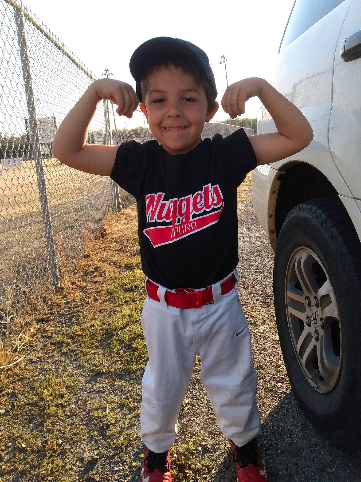 Mikey is registered to the contest to win money with this photo: alloy_wheel, automotive_lighting, automotive_tire, bumper, car, fender, happy, hood, joy, mammal, motor_vehicle, person, sky, sleeve, smile, t_shirt, tints_and_shades, tire, toddler, vehicle