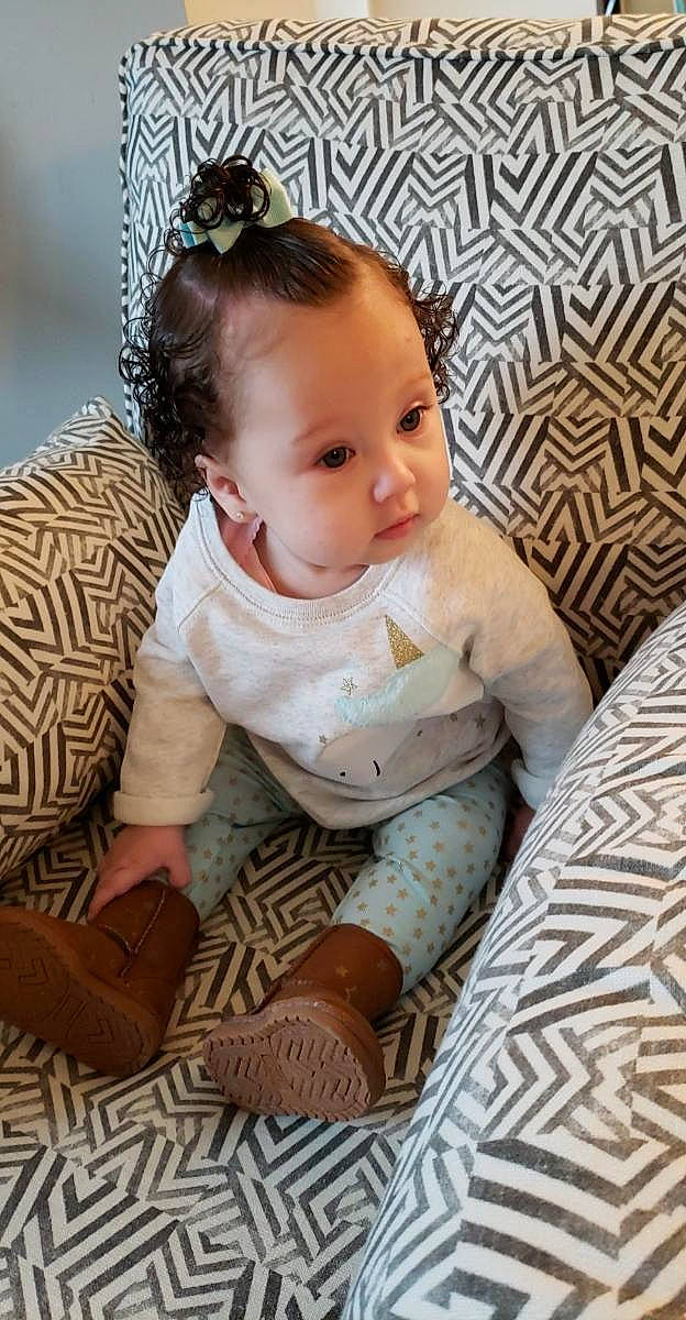 Gisella is registered to the contest to win money with this photo: arm, baby, baby_toddler_clothing, brown, cheek, child, ear, hair_accessory, headgear, pattern, person, photography, product, room, sitting, skin, toddler