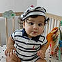 baby, child, crib, striped_clothing, hat, socks, indoor, cute, infant, face, curly_hair, person, sitting, nursery, toy, white_background, patterned_bedding, portrait, smile, young_child