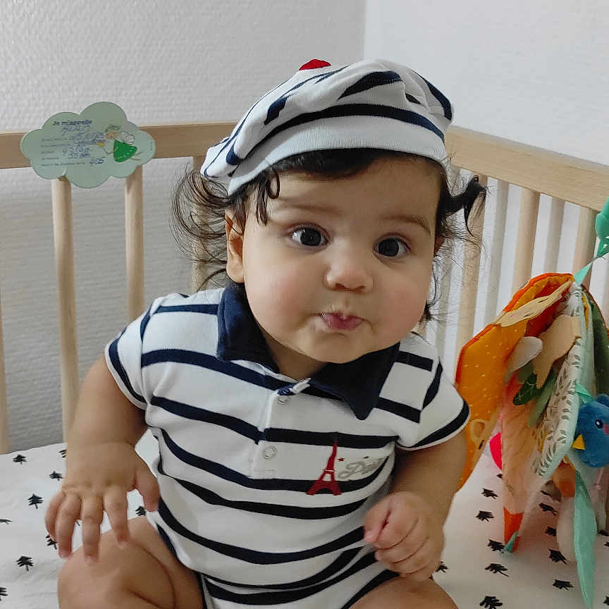 Adib participe au concours pour gagner de l'argent avec cette photo : baby, child, crib, curly_hair, cute, face, hat, indoor, infant, nursery, patterned_bedding, person, portrait, sitting, smile, socks, striped_clothing, toy, white_background, young_child