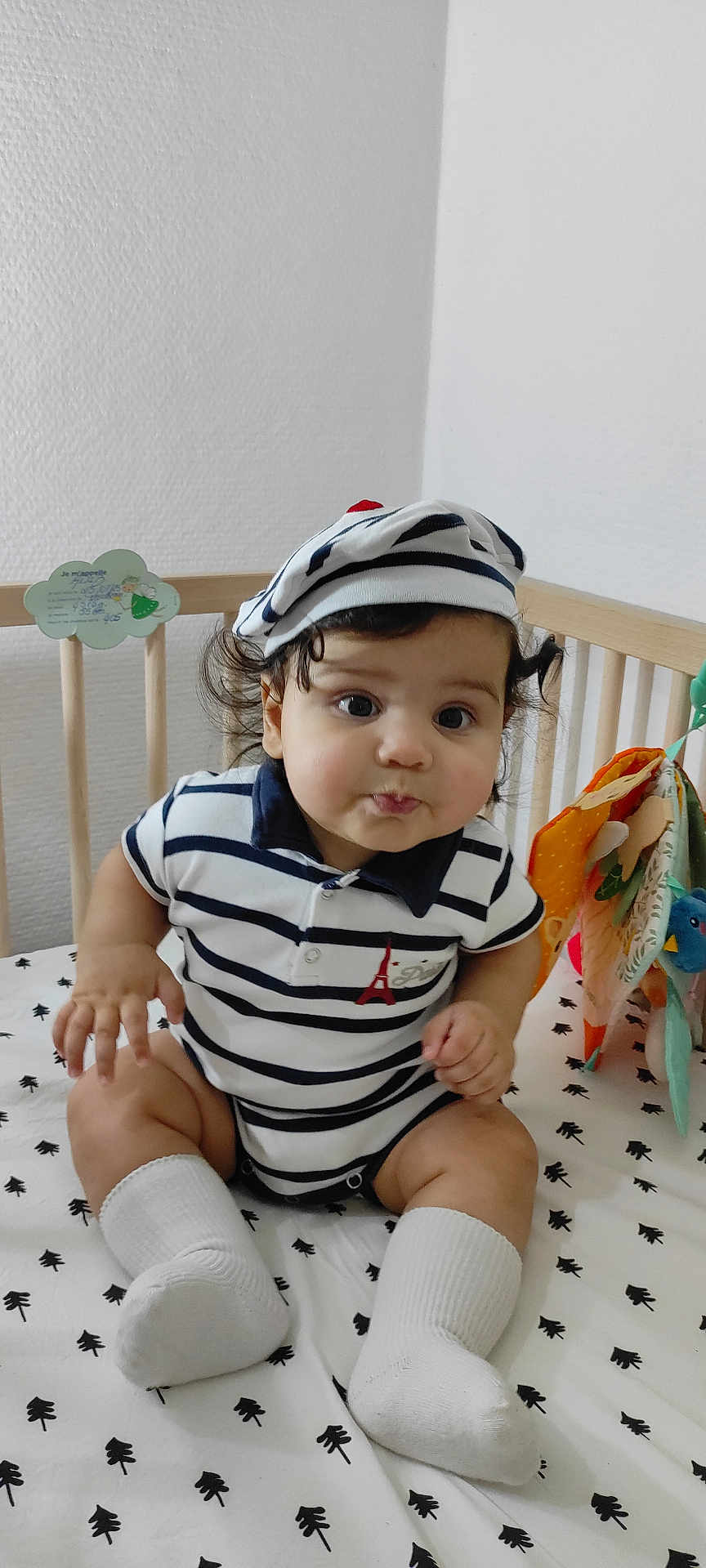 Adib participe au concours pour gagner de l'argent avec cette photo : baby, child, crib, striped_clothing, hat, socks, indoor, cute, infant, face, curly_hair, person, sitting, nursery, toy, white_background, patterned_bedding, portrait, smile, young_child