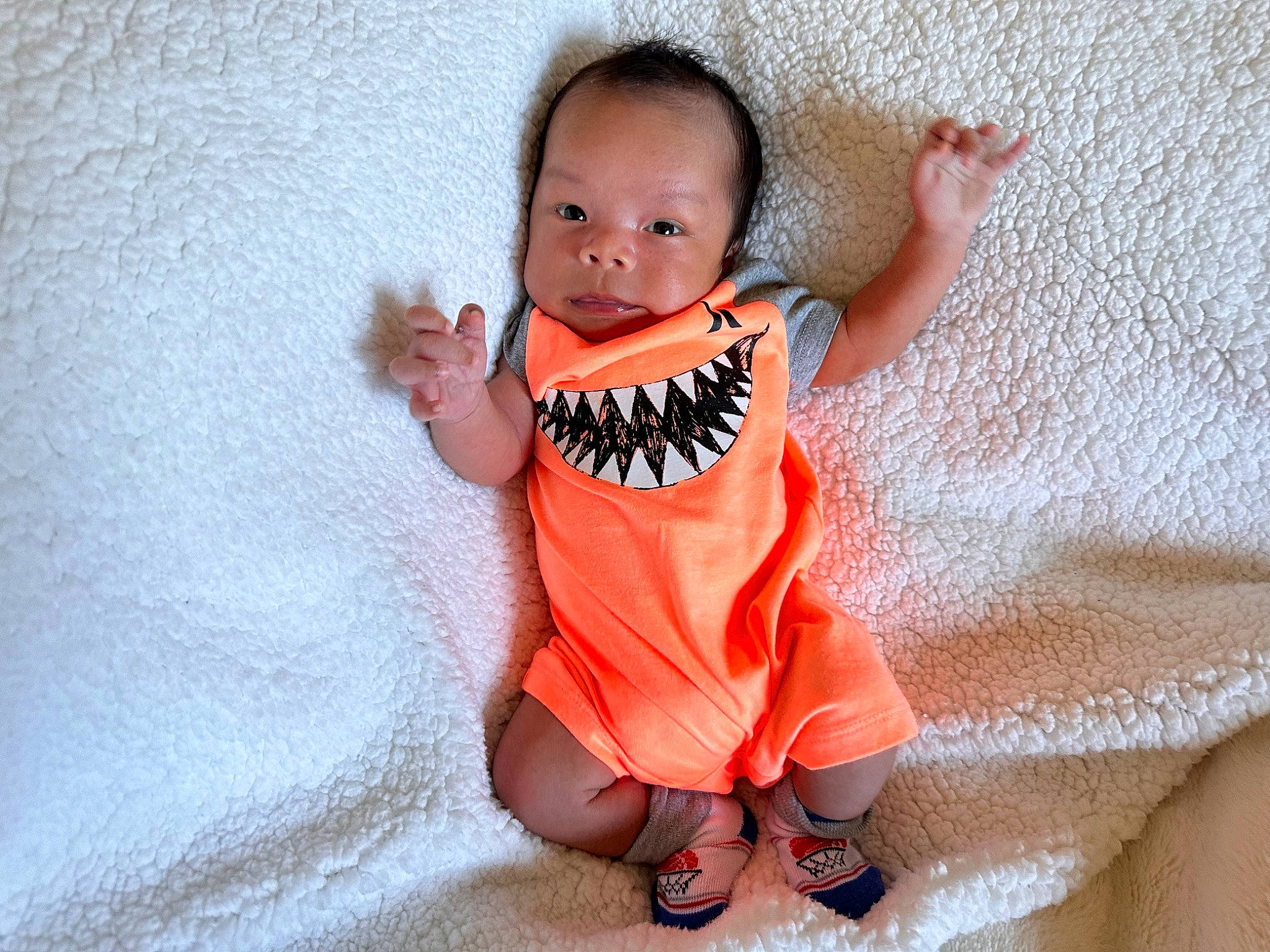 Sebastian is registered to the contest to win money with this photo: arm, baby, baby_toddler_clothing, cheek, child, eye, face, flooring, foot, gesture, happy, linens, orange, pattern, person, sitting, sleeve, t_shirt, thumb, toddler