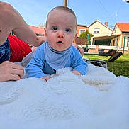 Jules a rejoint le concours — aidez-le/la à gagner de superbes lots ! baby, infant, blue_clothing, bib, blanket, outdoor, garden, grass, swing, adult_arm, house, sky, daylight, lawn, curious_expression, child, portrait, person, sunny, relaxing