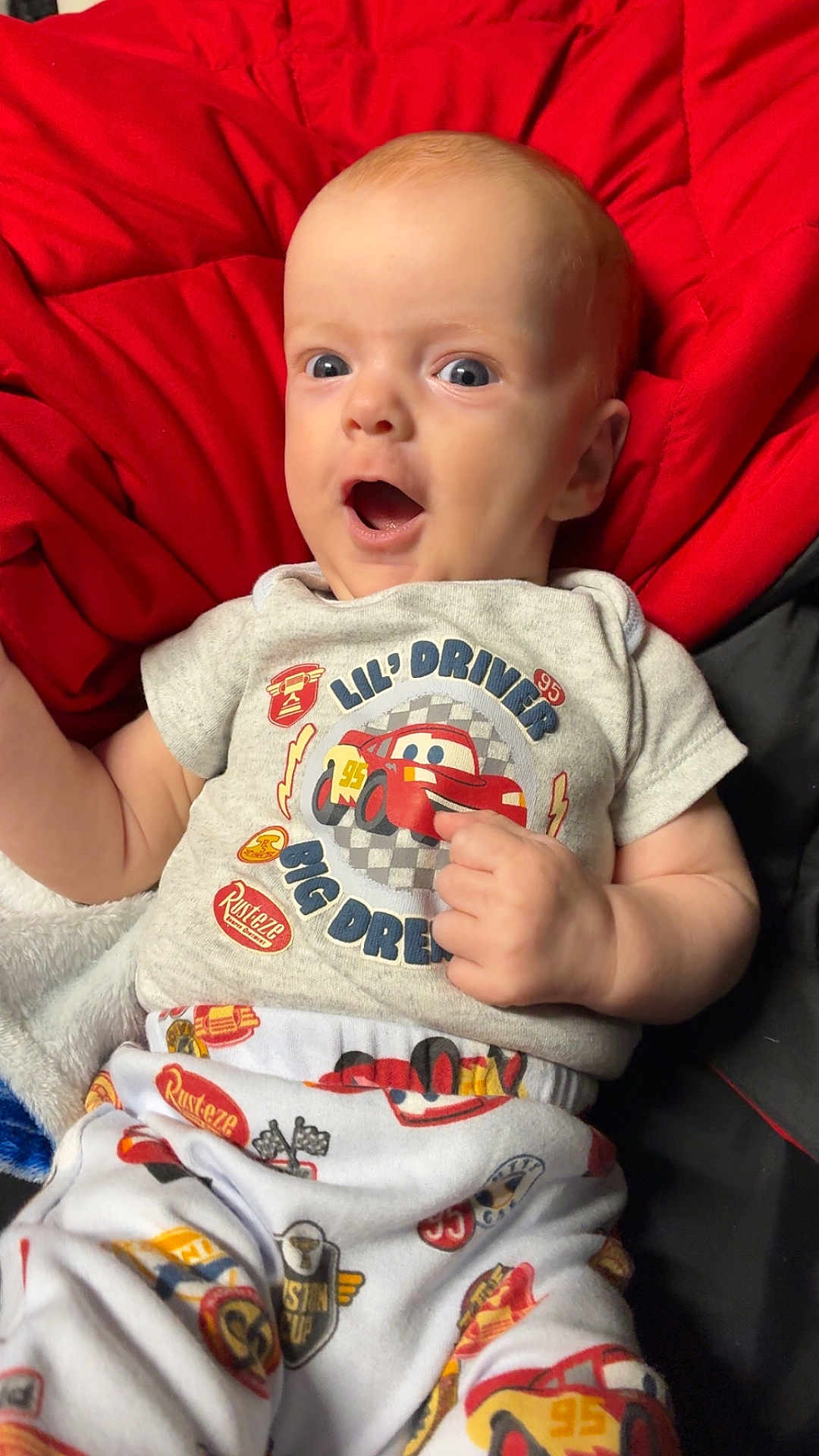 Atticus is registered to the contest to win money with this photo: baby, infant, child, clothing, red_blanket, white_pants, cute, surprised_expression, lying_down, cartoon_car, short_sleeve_shirt, indoor, person, face, hand, blue_eyes, light_skin, fabric, patterned_clothing, cozy