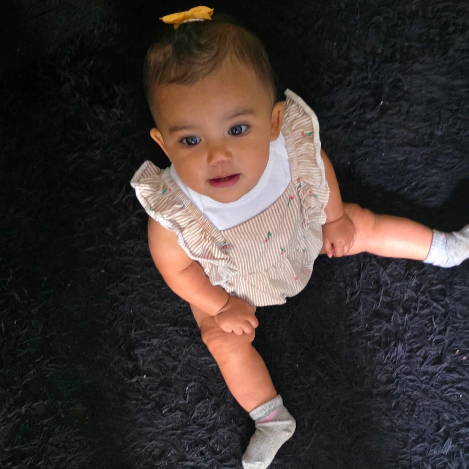 Stallace is registered to the contest to win money with this photo: baby, child, infant, sitting, dress, ruffles, yellow_bow, socks, black_rug, curious, looking_up, cute, indoors, person, portrait, legs, hands, happy, soft_texture, casual_clothing