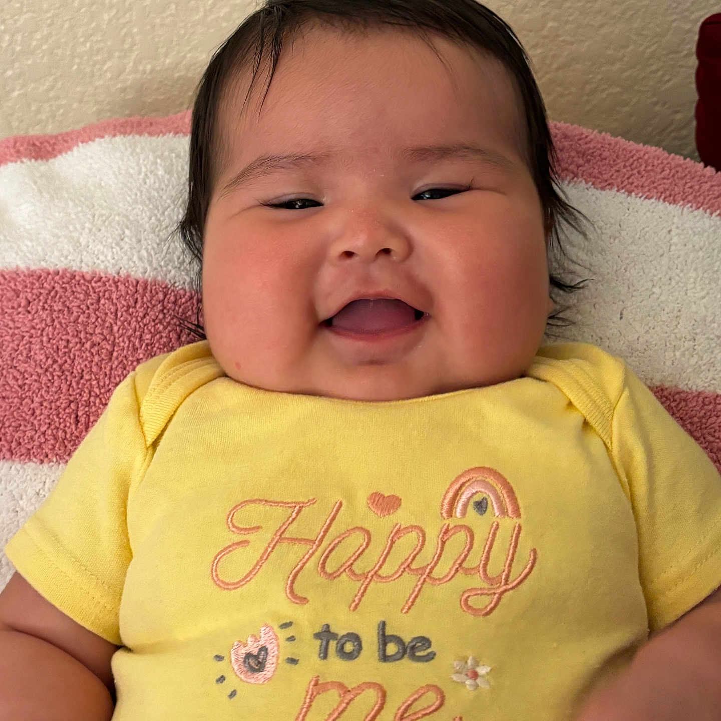 Jolene is registered to the contest to win money with this photo: baby, smiling, yellow_onesie, towel, pink, white, happy, embroidery, text_on_clothing, closeup, infant, face, chubby_cheeks, indoor, resting, cute, child, portrait, person, soft_texture