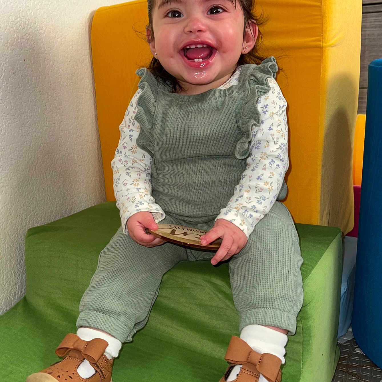 Edna is registered to the contest to win money with this photo: baby, brown_shoes, child, colorful, cushion, cute, furniture, green_outfit, happy, holding_object, indoor, plaque, playroom, portrait, short_hair, sitting, smile, step, toddler, white_socks