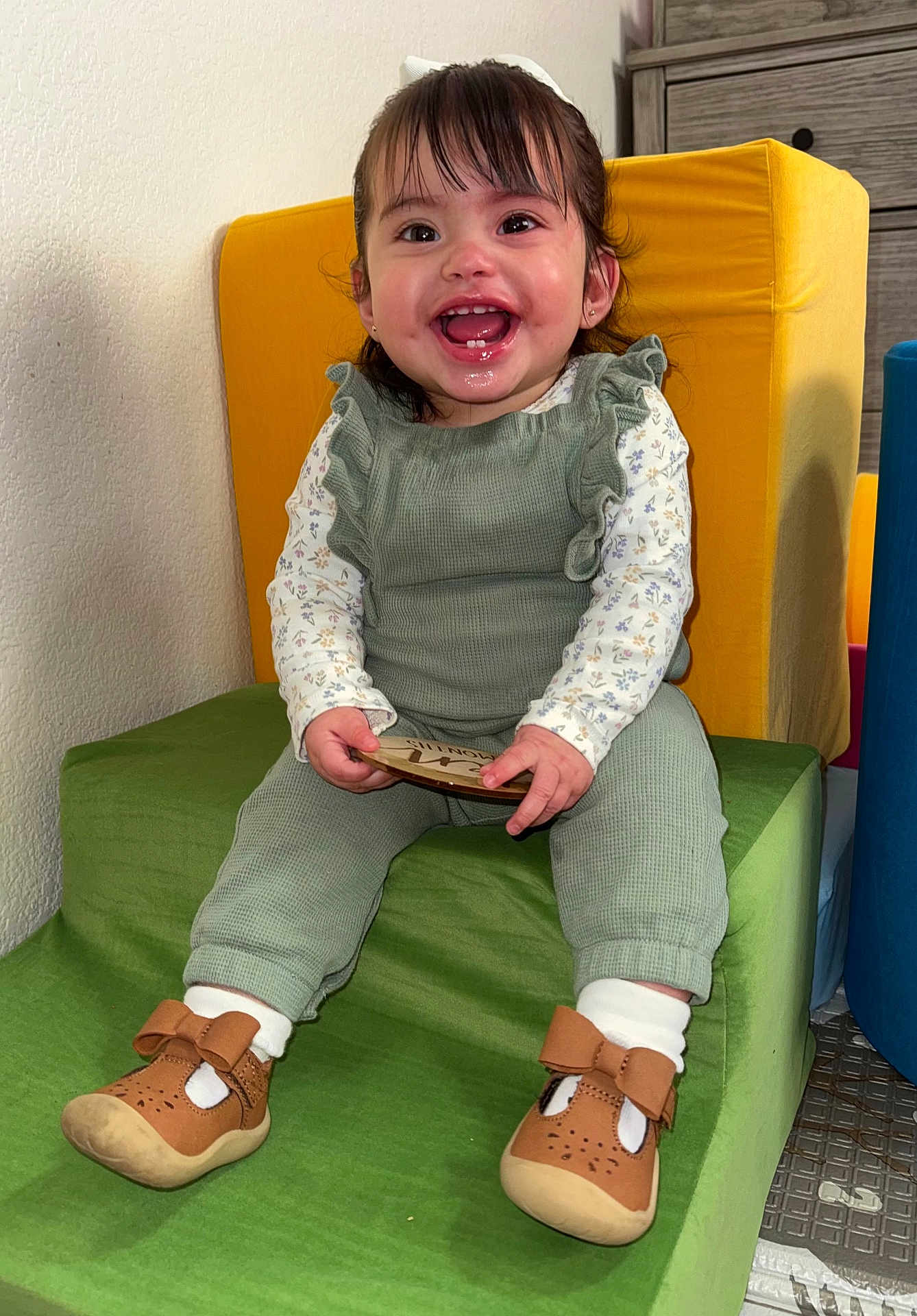 Edna is registered to the contest to win money with this photo: toddler, child, smile, green_outfit, brown_shoes, cushion, indoor, happy, holding_object, plaque, short_hair, white_socks, furniture, step, cute, portrait, baby, playroom, colorful, sitting