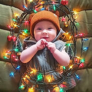 Jasmine is registered to the contest to win money with this photo: baby, child, infant, hat, bear_ears, vest, christmas_lights, holiday, festive, smiling, cute, colorful_lights, small_trees, quilted_background, indoor, portrait, happy, hands, warm_clothing, celebration