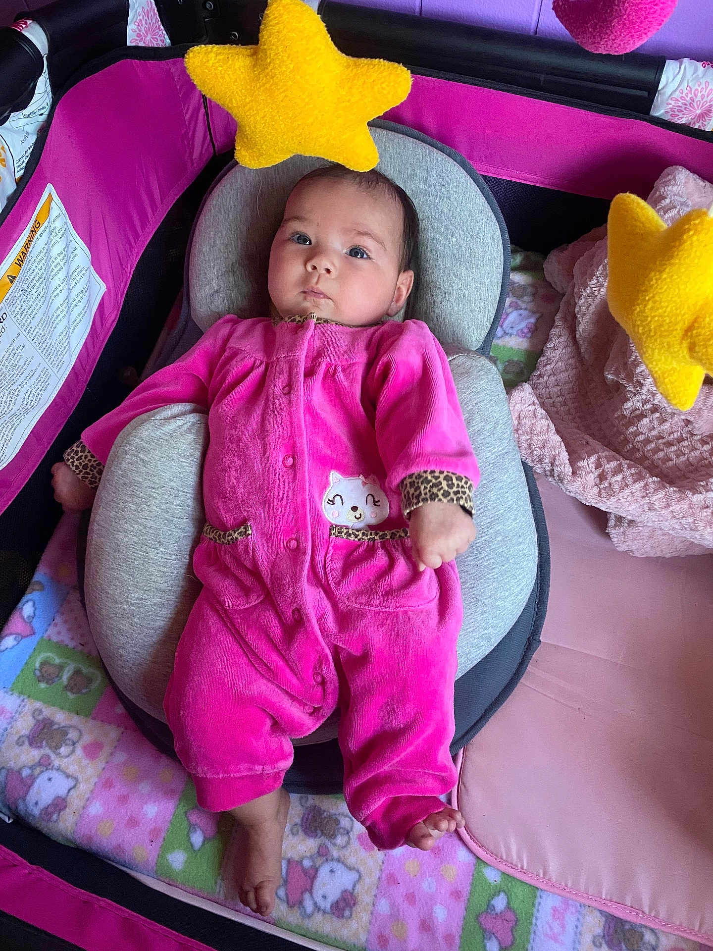 Zara is registered to the contest to win money with this photo: baby, infant, onesie, pink_clothing, baby_seat, cushion, playpen, yellow_star_toy, blanket, soft_toys, indoor, child, cute, face, feet, looking_up, comfort, resting, patterned_blanket, colorful