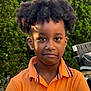 child, boy, outdoor, greenery, tree, face, portrait, expression, hair, orange_shirt, collar, casual_clothing, nature, daylight, person, young, standing, serious, candid, background