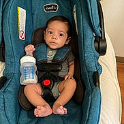 Juan joined the competition — help win amazing prizes! baby, infant, car_seat, bottle, teal, blue, seatbelt, child, indoors, cute, person, sitting, footwearless, wood_floor, wall, safety_harness, expression, looking_at_camera, young, casual_clothing