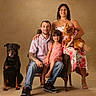 family, portrait, man, woman, child, girl, dog, rottweiler, small_dog, pet, chair, smiling, happy, casual_clothing, floral_dress, indoor, studio, seated, tattoo, footwear