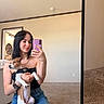 black_hair, carpet, casual_clothing, denim_skirt, dog, doorway, harness, holding, indoor, mirror, mirror_reflection, pet, seated, selfie, smartphone, smile, tattoo, technology, wall, woman