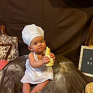 Anya is registered to the contest to win money with this photo: baby, chef_hat, apron, dough, bread, cutting_board, flour, bakery, sign, food, sitting, curious, kitchen, child, brown_background, table, colorful_bread, portrait, indoors, cute