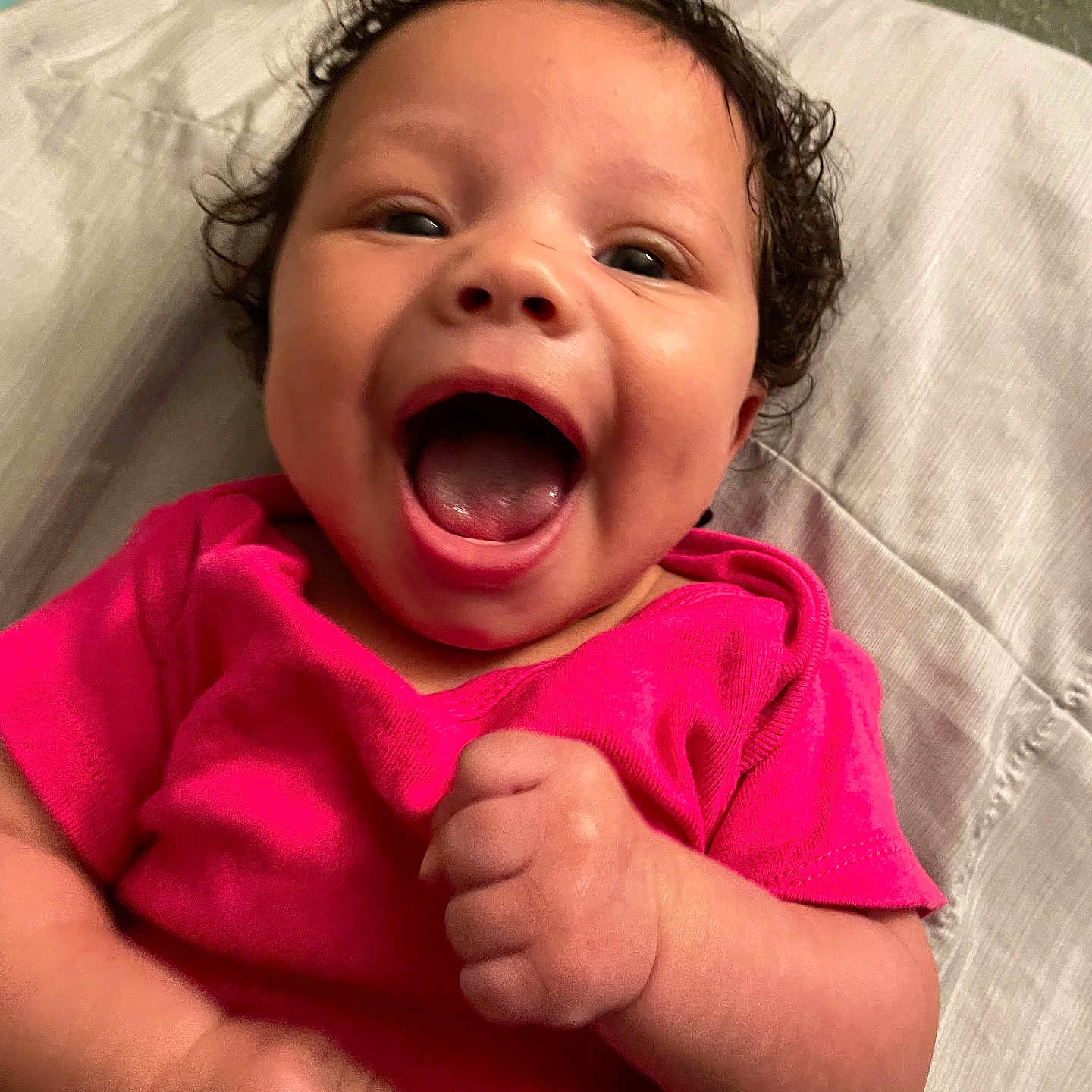 Remii is registered to the contest to win money with this photo: baby, child, closeup, curly_hair, cute, expression, face, hands, happy, indoors, infant, joy, laying_down, mouth_open, person, pink_shirt, portrait, skin, smiling, young_child