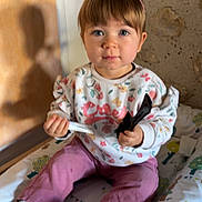 Lilas a rejoint le concours — aidez-le/la à gagner de superbes lots ! toddler, child, indoor, pink_bow, combed_hair, sitting, clothing, socks, face, curious, toy, padded_surface, patterned_blanket, stone_wall, light_shadow, person, holding, expression, young_child, casual_wear