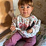 toddler, child, indoor, pink_bow, combed_hair, sitting, clothing, socks, face, curious, toy, padded_surface, patterned_blanket, stone_wall, light_shadow, person, holding, expression, young_child, casual_wear